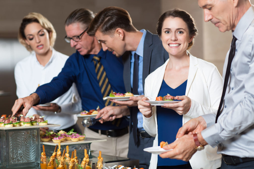 Smiling senior and middle-aged businessmen and businesswomen choosing snacks at buffet table. One of women is holding plate and smiling at camera.